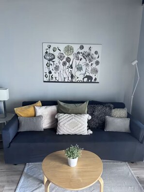 A modern living room with a dark blue sofa, multiple cushions, a round wooden coffee table, a small potted plant, and a wall-mounted abstract art piece. - Udacha this is a fully renovated studio (Montego Bay)