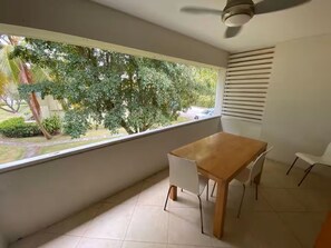 Ein Balkon mit Holztisch und -stühlen, einem Deckenventilator und Blick auf Bäume sowie ein Auto durch das Fenster.
