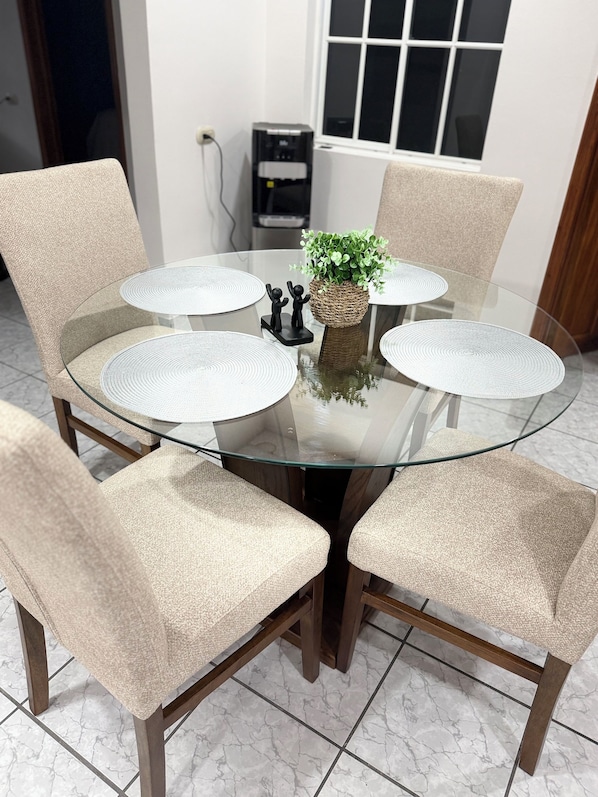 A round glass table with four chairs, a small plant, and a water dispenser in the background.