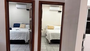 Un pasillo que conduce a dos dormitorios con camas blancas, una mesita y un aire acondicionado.