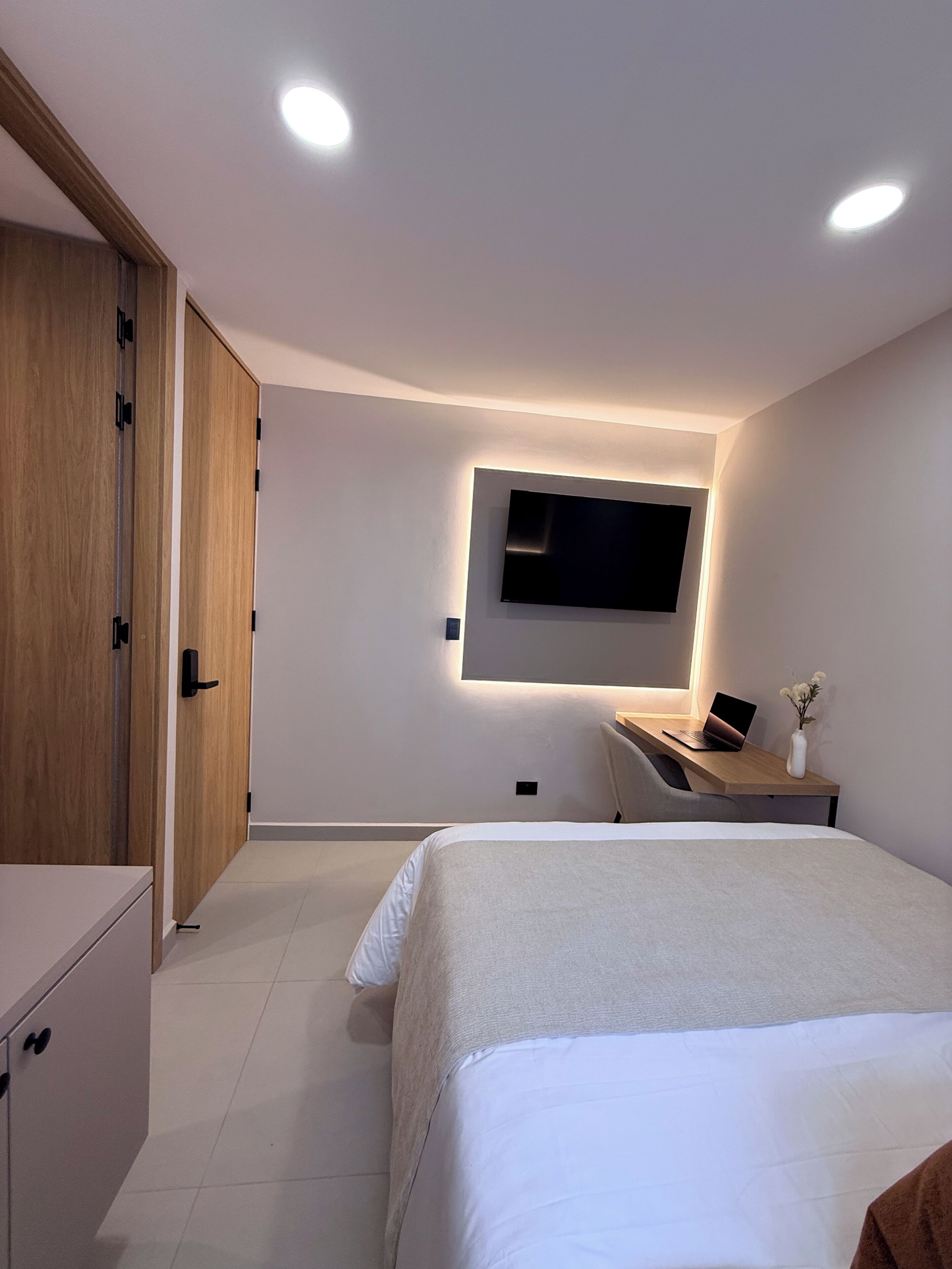 Quarto moderno com cama, escrivaninha com laptop, TV fixada na parede e guarda-roupa de madeira.