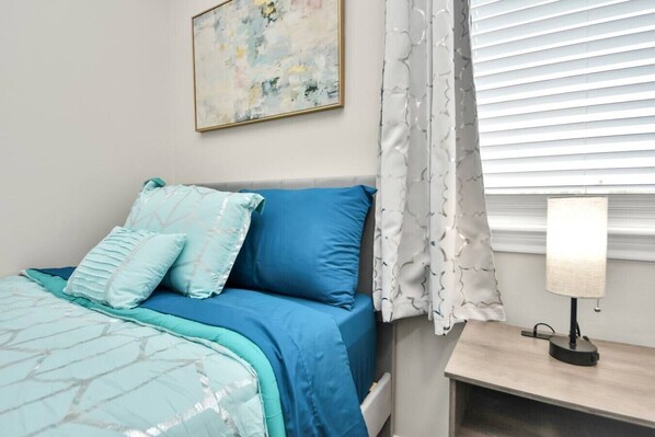 A bedroom with a bed, pillows, a bedside table with a lamp, a window with blinds, and a painting on the wall.