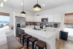 A modern kitchen with white cabinets, a central island with stools, a stainless steel refrigerator, a microwave, and a coffee maker. - ☀️Villas Laguna #2 - Exceptional Views, Pool, Walk to Beach (Panama City Beach)