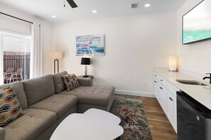 A modern living room with a sectional sofa, a white coffee table, a patterned rug, a painting on the wall, and a kitchen area with a sink and cabinets. - 🌊 Villas at Laguna Beach 3 - 5 BR, Beach, Pool, Rooftop Terrace (Panama City Beach)