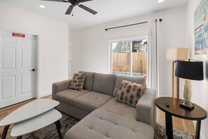 A modern living room with a sofa, two patterned cushions, a small table, a ceiling fan, and a window with a view of a wooden deck. - 🌊 Villas at Laguna Beach 3 - 5 BR, Beach, Pool, Rooftop Terrace (Panama City Beach)