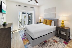Ein modernes Schlafzimmer mit einem großen Bett, zwei Nachttischen mit Lampen, einer Kommode, einem Deckenventilator und Blick ins Freie.