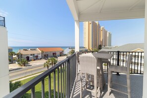 A balcony with wicker furniture overlooking a street and buildings, with a clear view of the ocean. - 🌊 Villas Laguna #8 - Beach Retreat, Pool, Rooftop Terrace, Pets! (Panama City Beach)