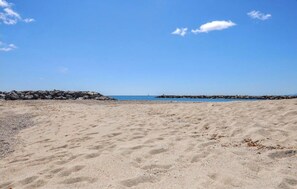 A sandy beach with a clear sky, a rocky breakwater, and calm water in the distance.