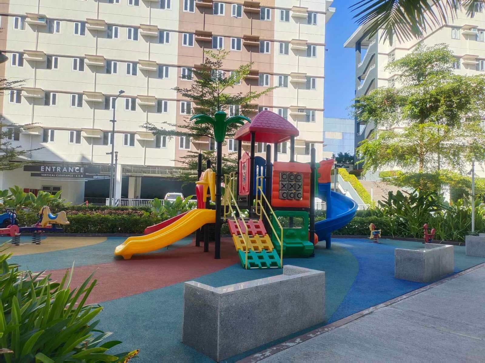 A playground with slides and climbing structures, surrounded by greenery and a multi-story building in the background.