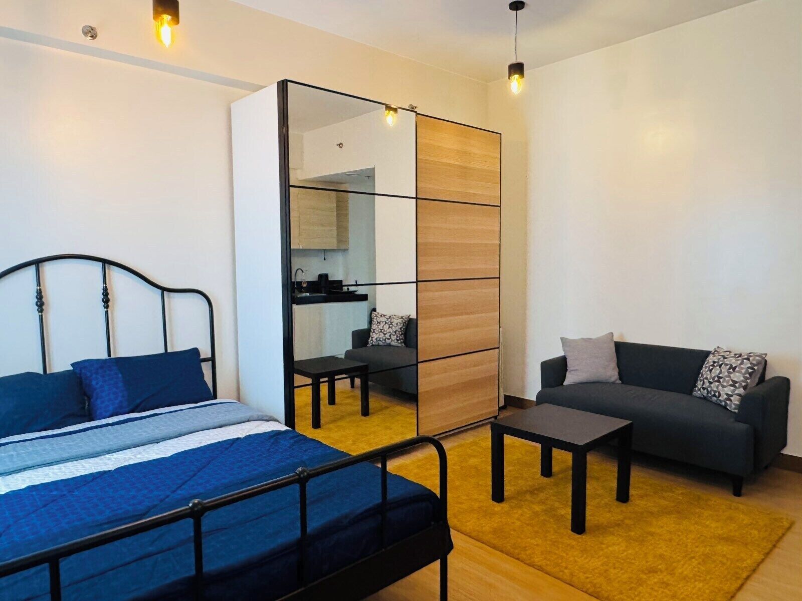 A bedroom with a bed, a sofa, a small table, a mirror, and a kitchenette.