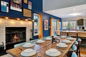 A dining area with a fireplace, a long wooden table set for six, and a kitchen visible through an open door. - 5BR Game Room, Hot Tub, Fire Pit + Lake Access (East Stroudsburg)