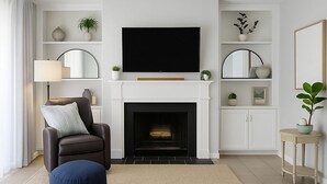 Ein Wohnzimmer mit Kamin, Fernseher, einer Couch, einem Sessel, einem runden Hocker und einem kleinen Tisch mit Pflanze.