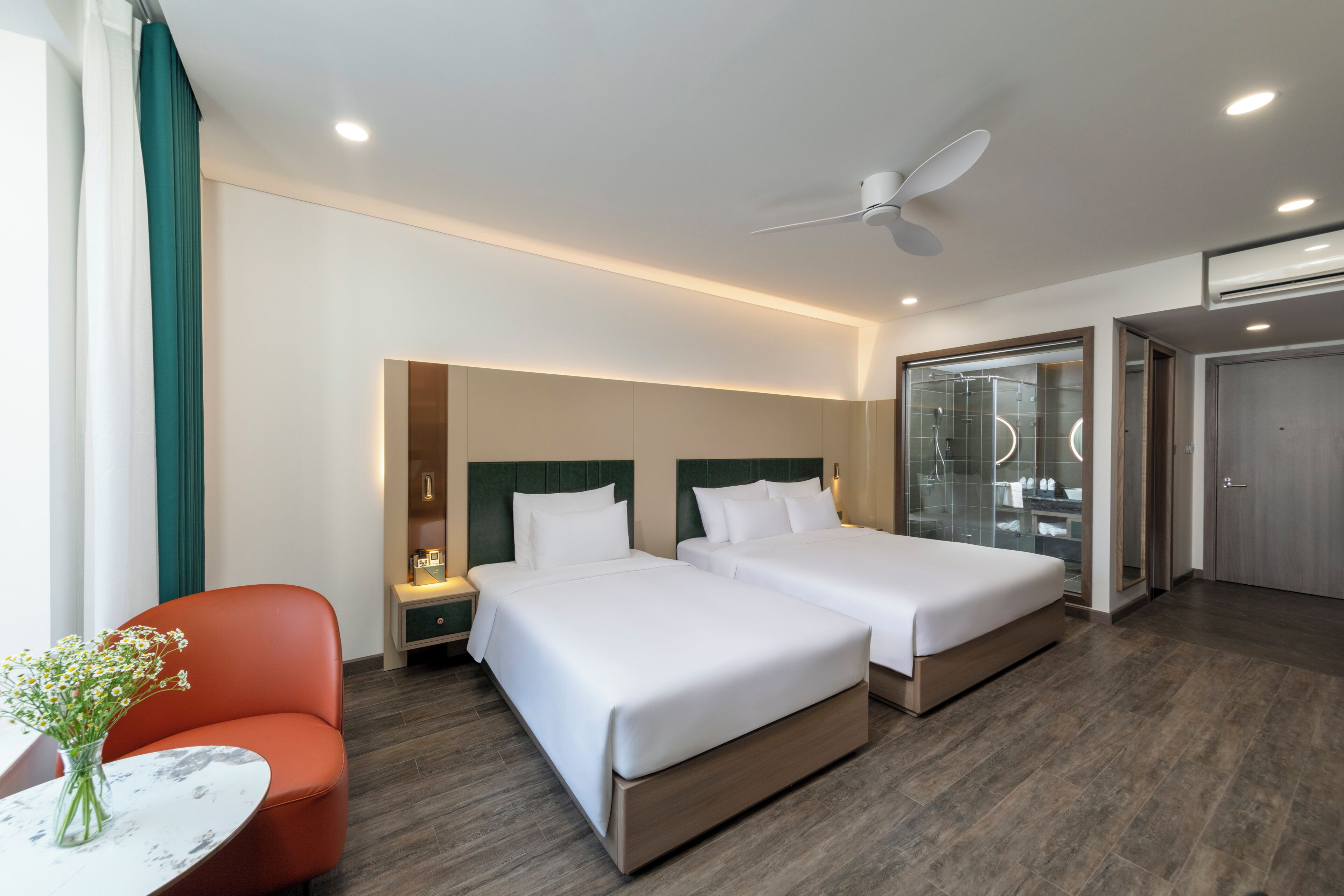 A hotel room with two beds, a nightstand, a red chair, a small table with a vase, and a bathroom with a glass shower enclosure.