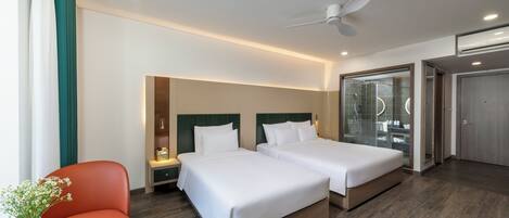 A hotel room with two beds, a nightstand, a red chair, a small table with a vase, and a bathroom with a glass shower enclosure.