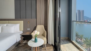 Hotellihuone, jossa on sänky, sohva, pieni pöytä maljakolla sekä näkymä kaupunkimaisemaan ja vesistöön.