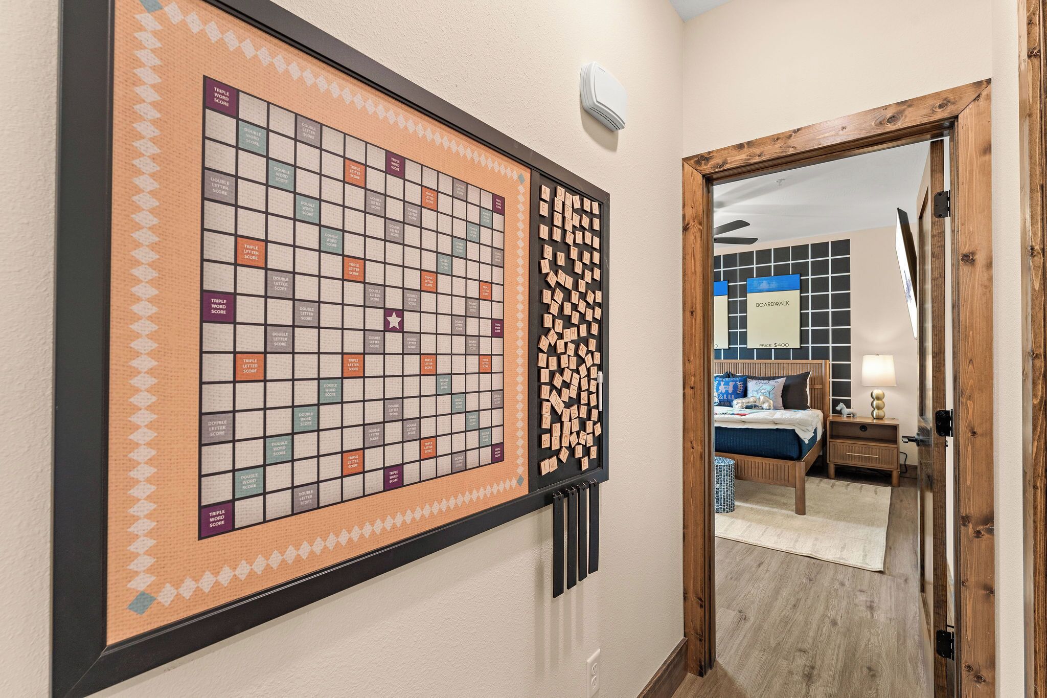 Une pièce avec un grand jeu de société accroché au mur, un lit recouvert d’une couverture bleue et une porte en bois.