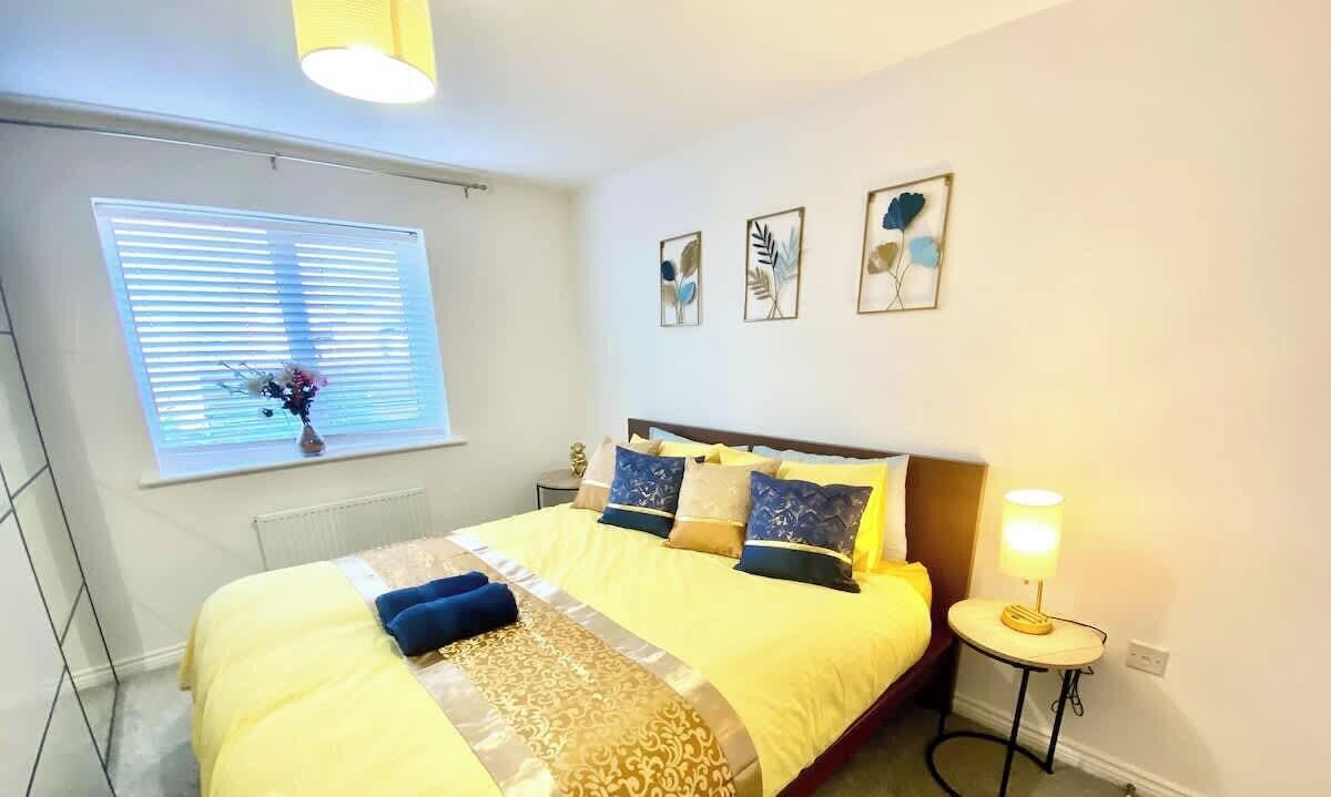 Cama bem arrumada com colcha amarela e almofadas decorativas, mesa de cabeceira com abajur e janela com persianas.