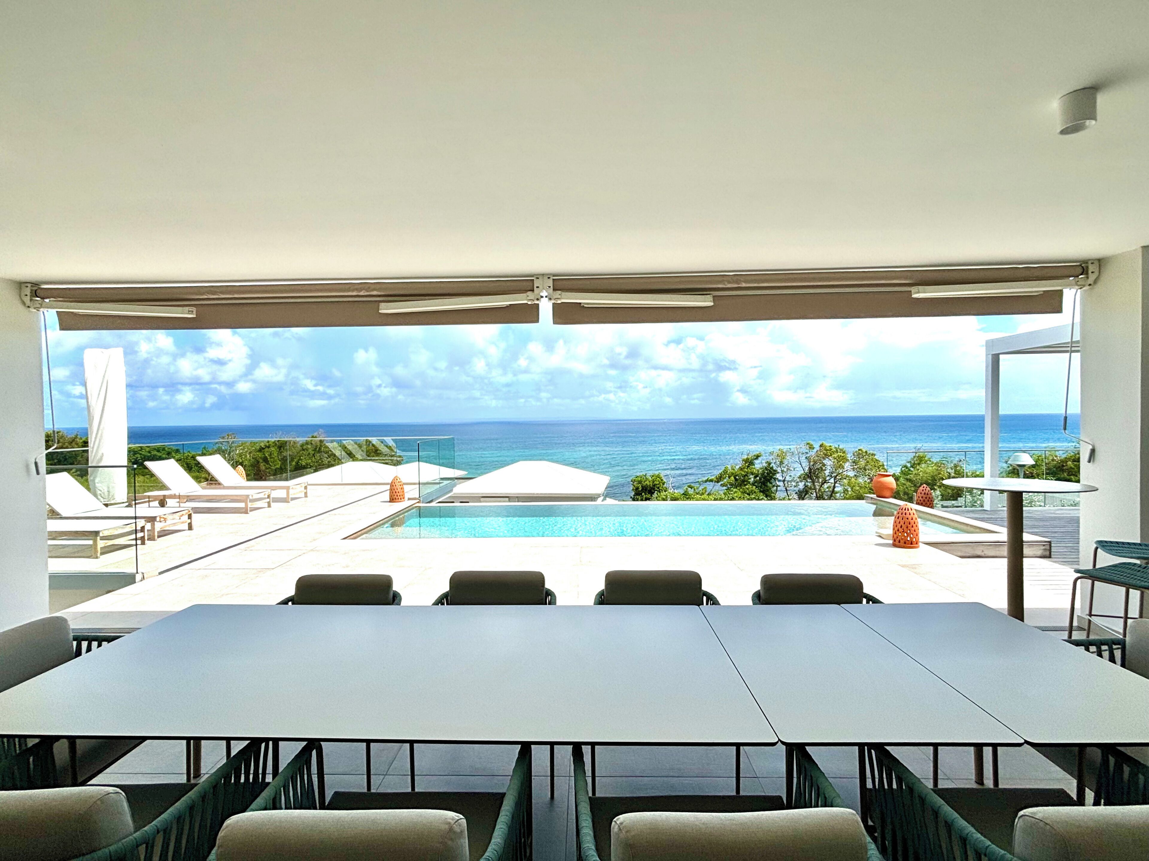 A modern outdoor pool area with lounge chairs, a dining table, and a view of the ocean.