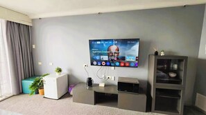 Un salon moderne avec un téléviseur à écran plat fixé au mur, un petit meuble blanc, un pouf turquoise et une plante en pot.