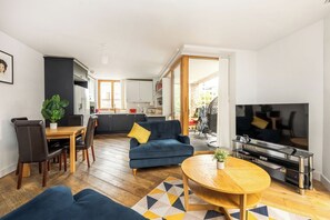 A modern living room with a sofa, coffee table, and TV. There is a dining area with chairs and a table, and a kitchen in the background. - Cool 2BR Apartment with Air Conditioning! (London)