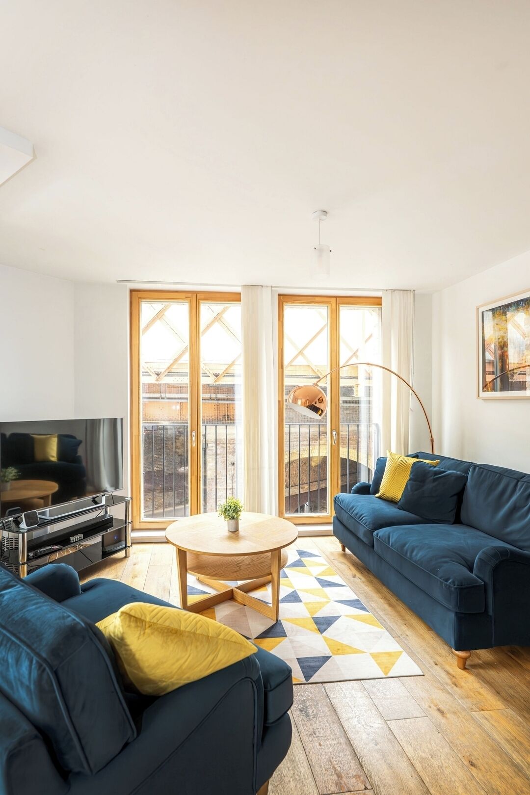En moderne stue med TV, blå sofa, gule puder, et sofabord og udsigt gennem store glasdøre.
