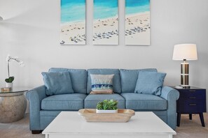 Een kamer met een blauwe bank, een witte salontafel, een bijzettafeltje met een lamp en een wand met drie strandafbeeldingen.