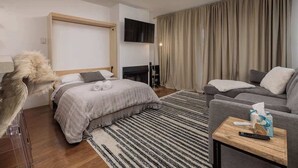A modern hotel room with a bed, sofa, TV, and curtains. - Ski-In Studio - Queen Murphy Bed - Walk to Lift (Girdwood)