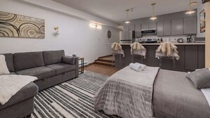 A modern apartment with a sofa, a bed, a kitchen area, and a staircase. - Ski-In Studio - Queen Murphy Bed - Walk to Lift (Girdwood)