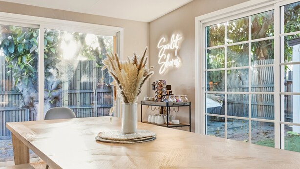 Un espace repas avec une table en bois, un vase contenant des plumes d’herbe des pampas et un panneau lumineux néon.