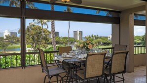 A dining area with a table set for four, large windows with a view of palm trees and buildings, and a ceiling fan. - Kaanapali Royal | Ocean View 4 BR, Sleeps 11 | Car Incl. w/ 6+ Nights | KRO ML-3794 by KBM (Lahaina)
