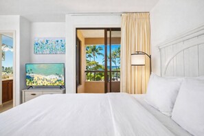 A hotel room with a bed, a television, a painting, a window with a view of palm trees, and a balcony. - Kaanapali Royal | Ocean View 6 BR, Sleeps 19 | Car Incl. w/ 6+ Nights | KRO ML-3797 by KBM (Lahaina)