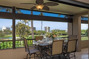 A dining area with a table set for four, large windows with a view of palm trees and buildings, and a ceiling fan. - Kaanapali Royal | Ocean View 6 BR, Sleeps 19 | Car Incl. w/ 6+ Nights | KRO ML-3797 by KBM (Lahaina)