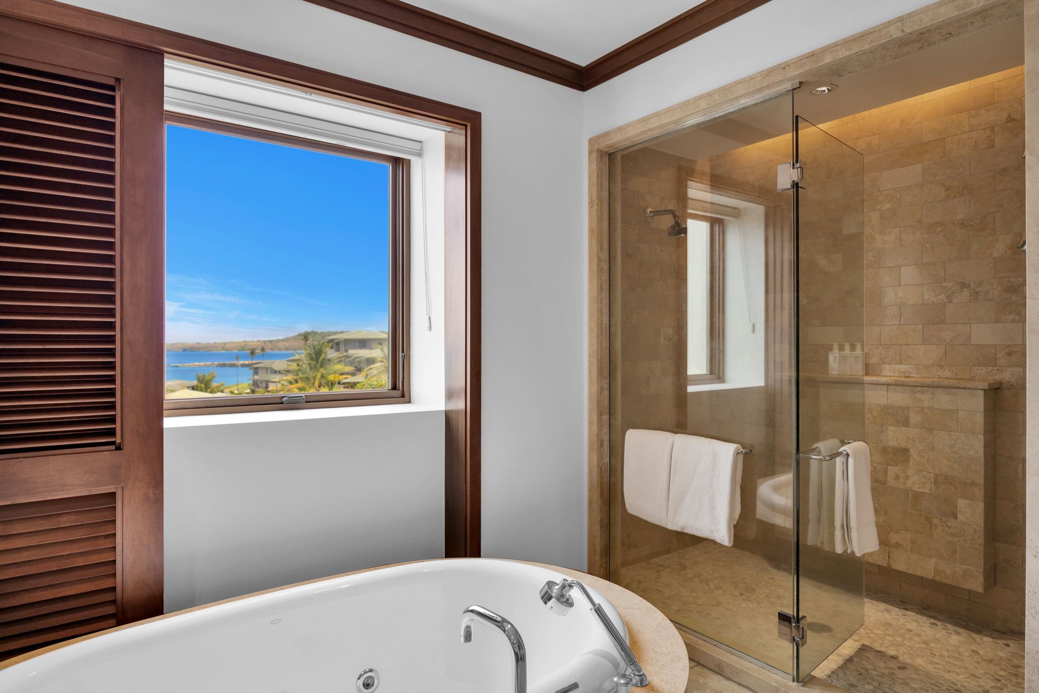 A bathroom with a bathtub, a glass-enclosed shower, and a window with a view of the beach.