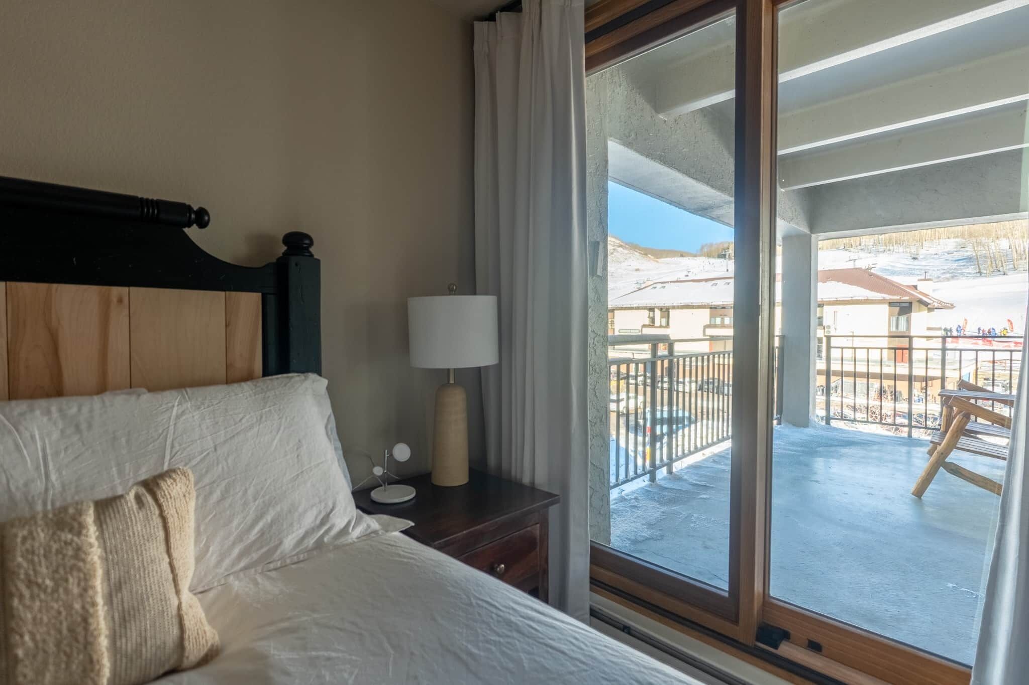 A hotel room with a bed, a nightstand, a lamp, and a large window with a view of a snowy outdoor area.