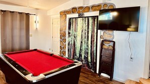 A pool table in a room with a stone wall, a television mounted on the wall, and a door with a forest mural.