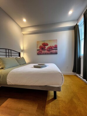 Een slaapkamer met een bed, een schilderij, een raam met gordijnen en een vloerbedekking.