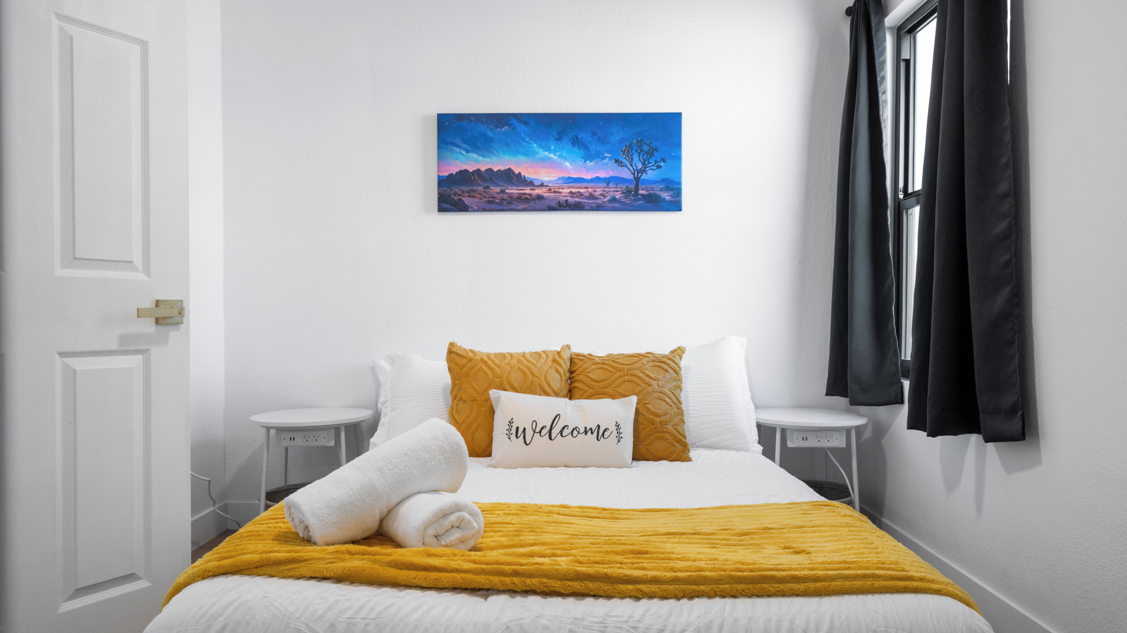 A neatly made bed with a yellow blanket, white pillows, and a 'Welcome' pillow, a painting of a landscape on the wall, and a window with black curtains.