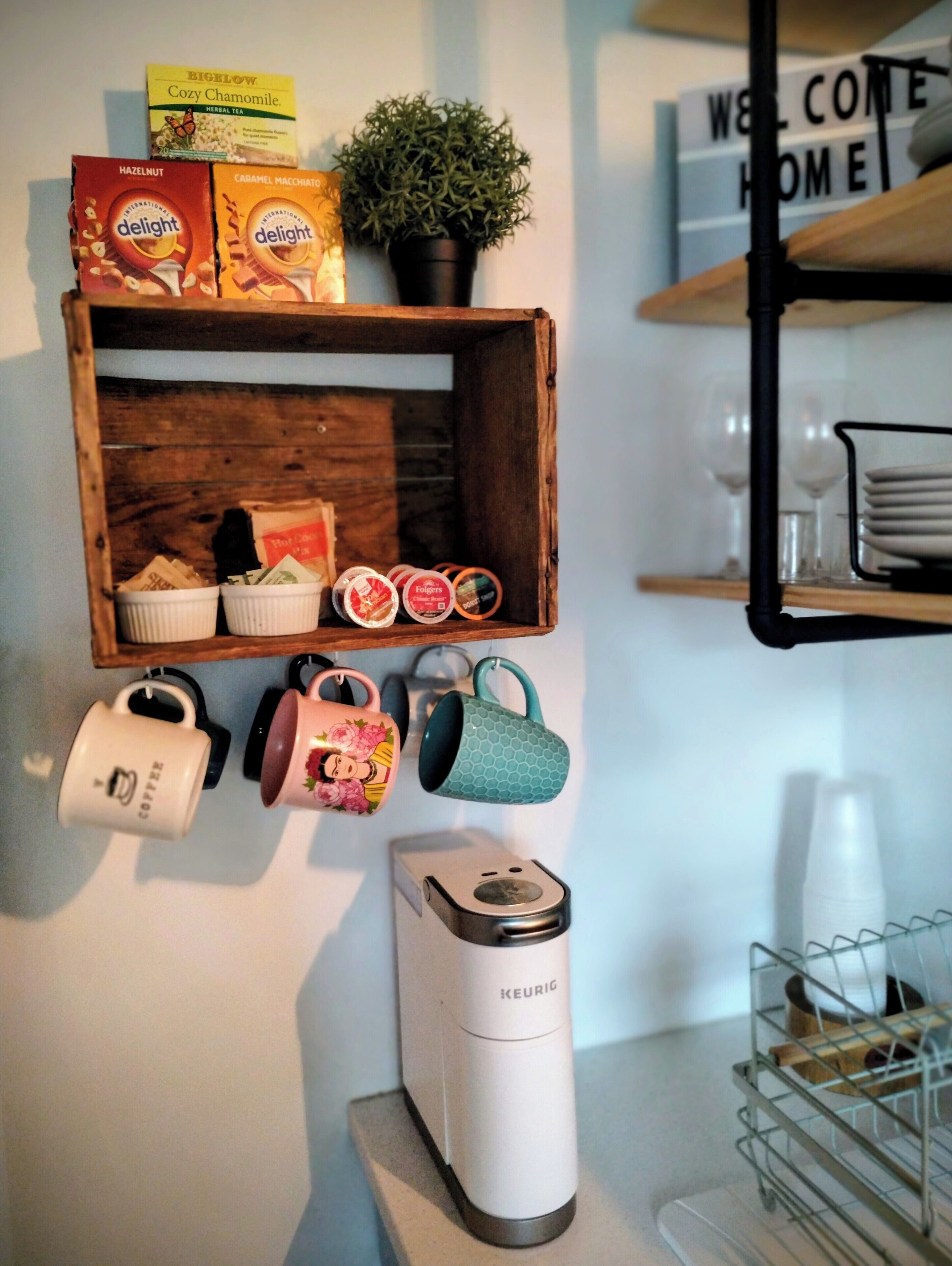 Un coin cuisine équipé d’une machine à café, de tasses, d’une étagère avec des boîtes de thé et d’une pancarte de bienvenue.