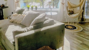 A room with a bed, a hammock, a window with curtains, a small table, and a basket.