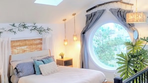 A bedroom with a skylight, a round window, a bed with pillows, a nightstand, a hanging light fixture, and a plant.