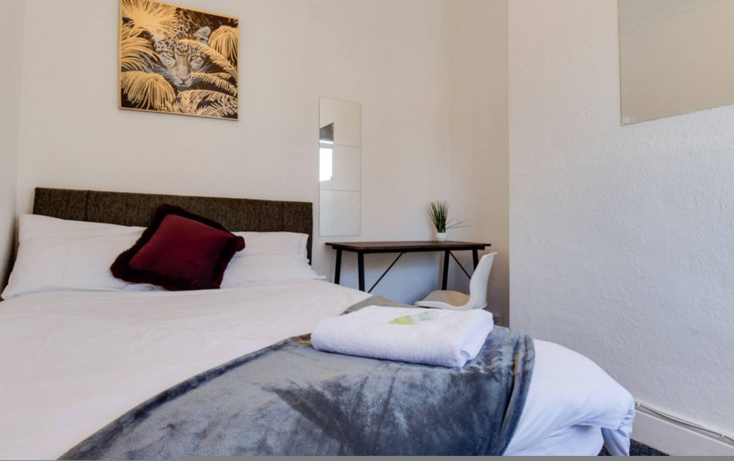 A hotel room with a bed, a red pillow, a small table with a plant, and a painting of a tiger on the wall.