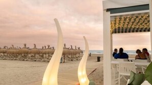 Un restaurante frente a la playa con terraza, vista al océano y sombrillas de paja de techo al fondo.