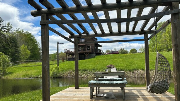 A wooden deck with a bench and a hanging chair, overlooking a body of water and a grassy hill with a house in the background.