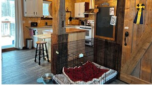 A rustic cabin interior with wooden walls, a kitchen area, a dining table, and a pet enclosure.