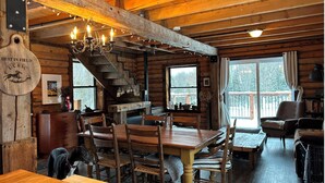 A rustic cabin interior with a wooden dining table, chairs, a staircase, and a chandelier.