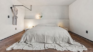Ein schlicht eingerichtetes Schlafzimmer mit einem großen Bett, einem Bademantel an einem Haken und einer Wandleuchte, sowie einer kleinen Pflanze.