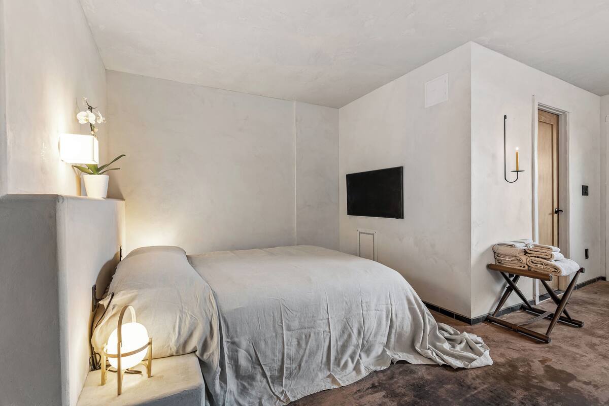 A bedroom with a bed, a bedside lamp, a wall-mounted TV, a door, and a small table with towels.