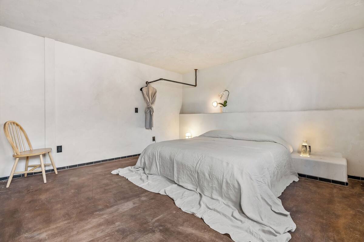 A bedroom with a bed, a chair, bedside tables, and a wall-mounted light fixture.