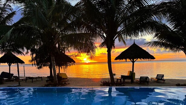 A poolside view with a sunset over the ocean, palm trees, and lounge chairs.
