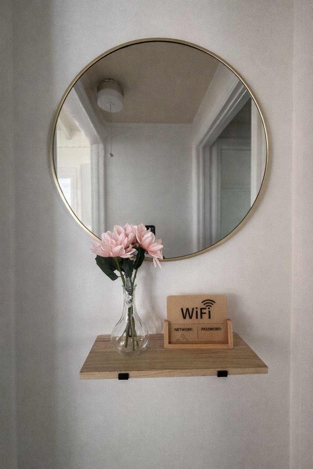 A mirror with a gold frame, a vase with pink flowers, and a wooden signboard with WiFi details.